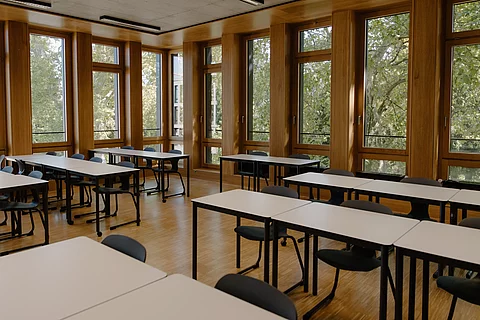 Das Foto zeigt ein leeres Klassenzimmer. Die Tische stehen in Reihen. Im Hintergrund sieht man eine große Fensterfront, durch die grüne Bäume zu sehen sind.