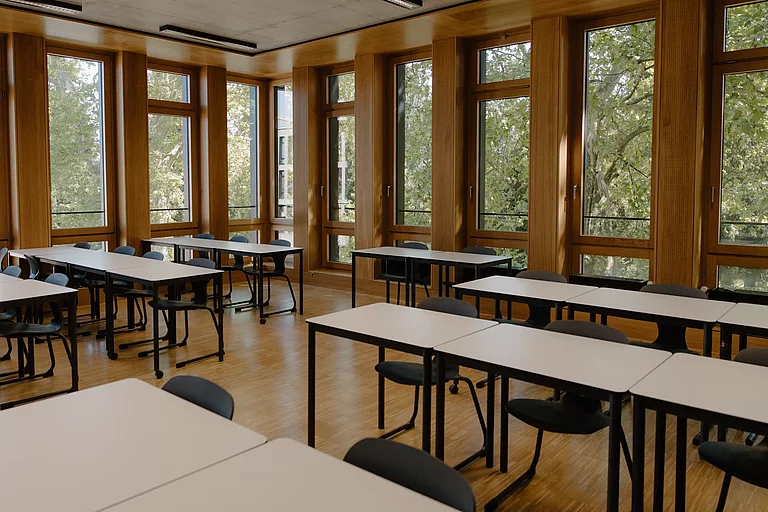 Das Foto zeigt ein leeres Klassenzimmer. Die Tische stehen in Reihen. Im Hintergrund sieht man eine große Fensterfront, durch die grüne Bäume zu sehen sind.