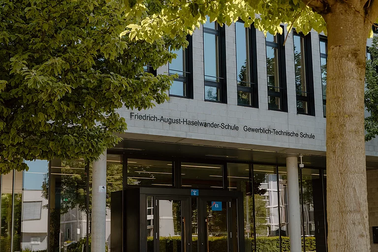 Das Foto zeigt den Haupteingang des G-Gebäudes. Der Bildausschnitt liegt auf dem Schriftzug über dem Eingang: Friedrich-August-Haselwander-Schule Gewerblich-Technische Schule.